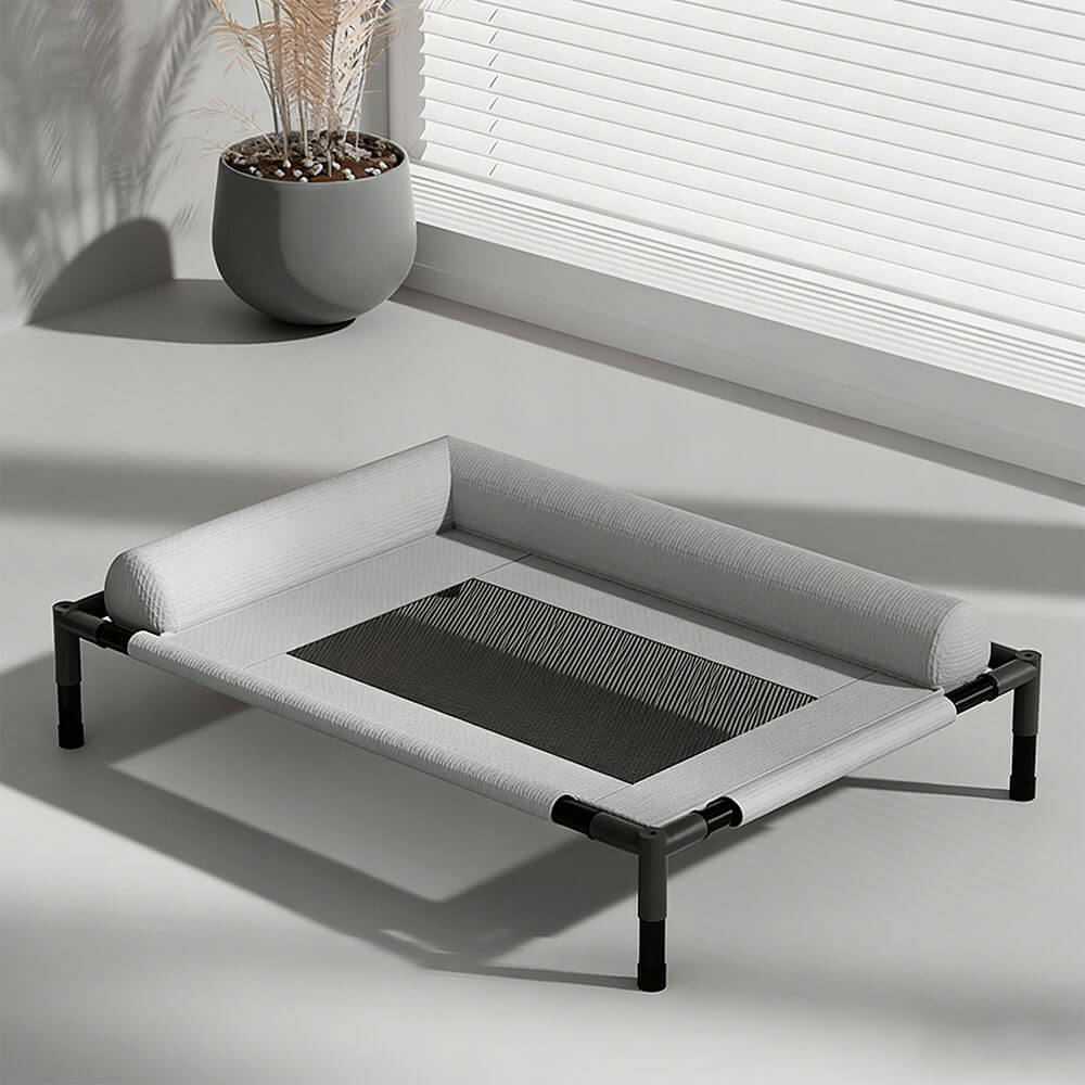 Raised pet bed with breathable mesh sleeping surface and soft bolster edges, placed near a window with blinds and a potted plant, ideal for indoor and outdoor use for dogs and cats.