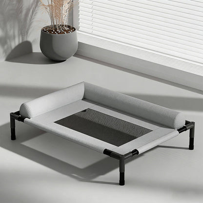 Raised pet bed with breathable mesh sleeping surface and soft bolster edges, placed near a window with blinds and a potted plant, ideal for indoor and outdoor use for dogs and cats.